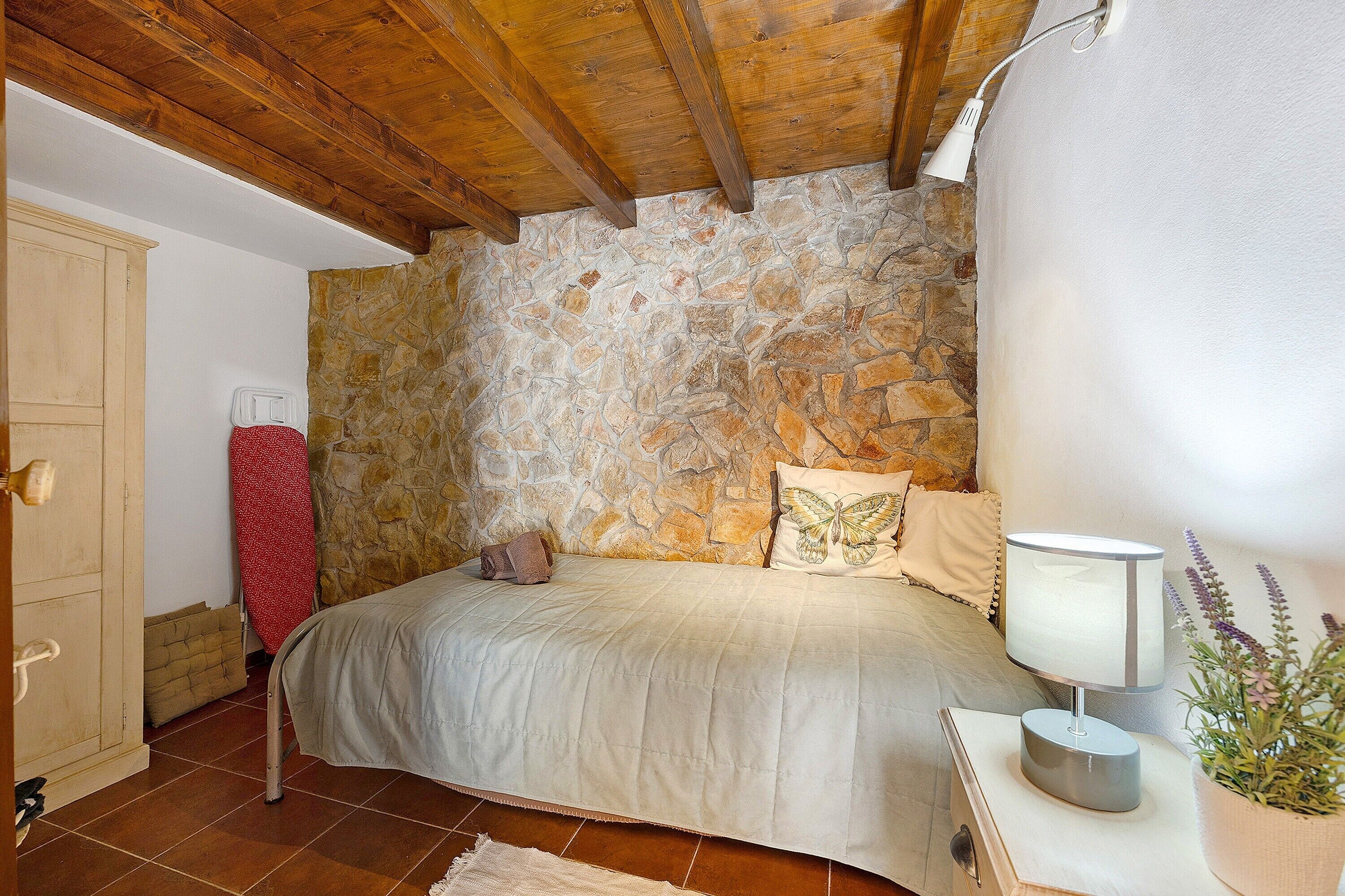 A bedroom with a stone wall, wooden ceiling beams, a bed with a white bedspread, a bedside lamp, and a potted plant.