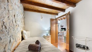Ein Schlafzimmer mit Steinwand, Holzbalkendecke, einem Bett mit Decke, einem Nachttisch und einem weißen Metallregal.