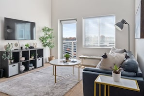 Ein modernes Wohnzimmer mit Flachbildfernseher, rundem Couchtisch, einer Couch, einer Stehlampe, einem Bücherregal und einer Topfpflanze.