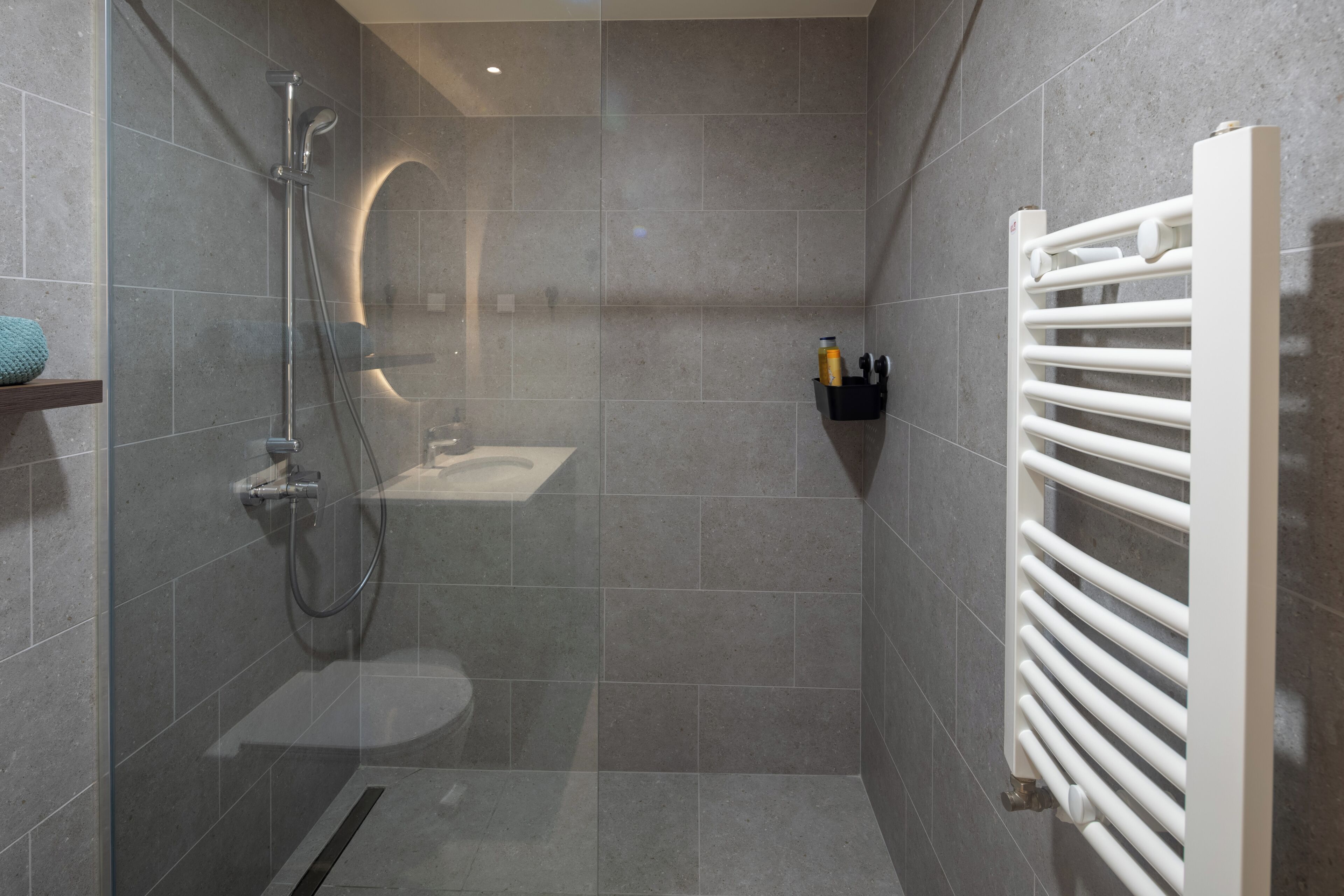 Une salle de bain moderne avec une douche à l’italienne en verre, un radiateur blanc et une petite étagère contenant des articles de toilette.