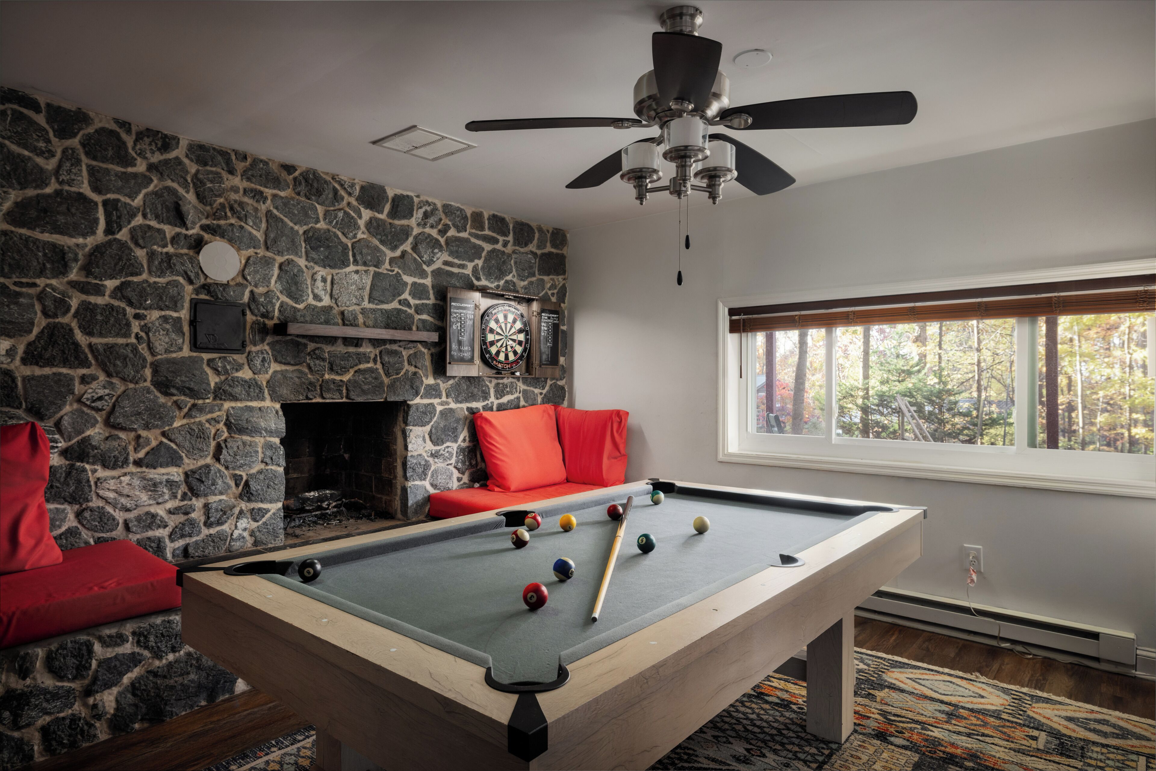 Une table de billard dans une pièce aux murs de pierre, avec une cheminée et une fenêtre donnant sur des arbres.