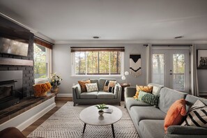 A living room with a fireplace, two sofas, a coffee table, and a large window with a view of trees. - Huckleberry Hills | Hot Tub & Fire Pit & Dogs (Harpers Ferry)