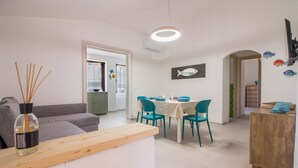 A modern living room with a sofa, dining table, chairs, and a wooden side table. - GuestHost - Cozy and bright 70 square meters apartment, perfect for four people, located on the first floor of a building without an elevator.The property enjoys an excellent location, in an extremely quiet area and just 250 meters from the seafront. Ame (Cannigione)