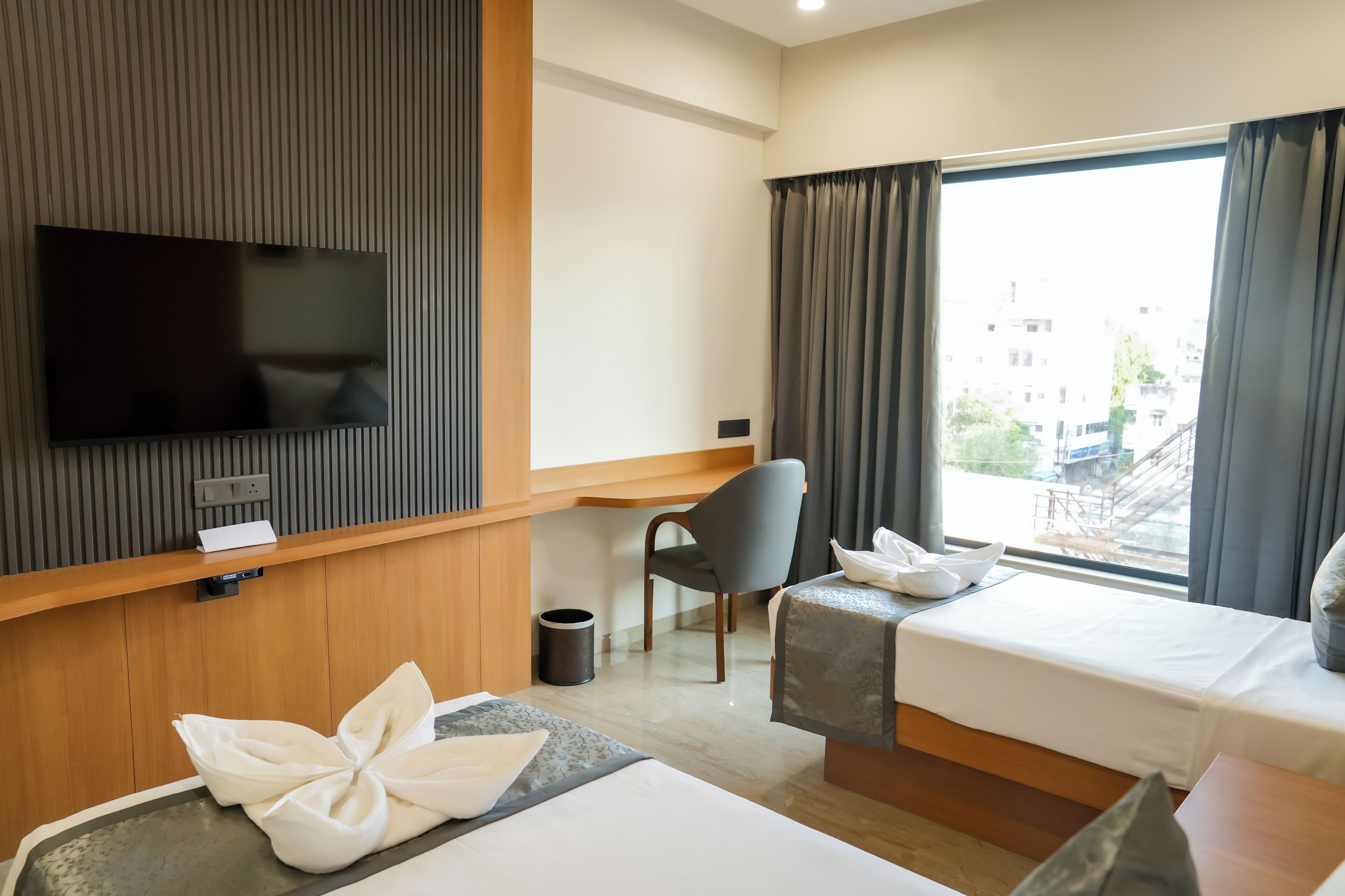 Quarto de hotel com TV de tela plana, escrivaninha, cadeira, cama, mesa de cabeceira e janela com cortinas.