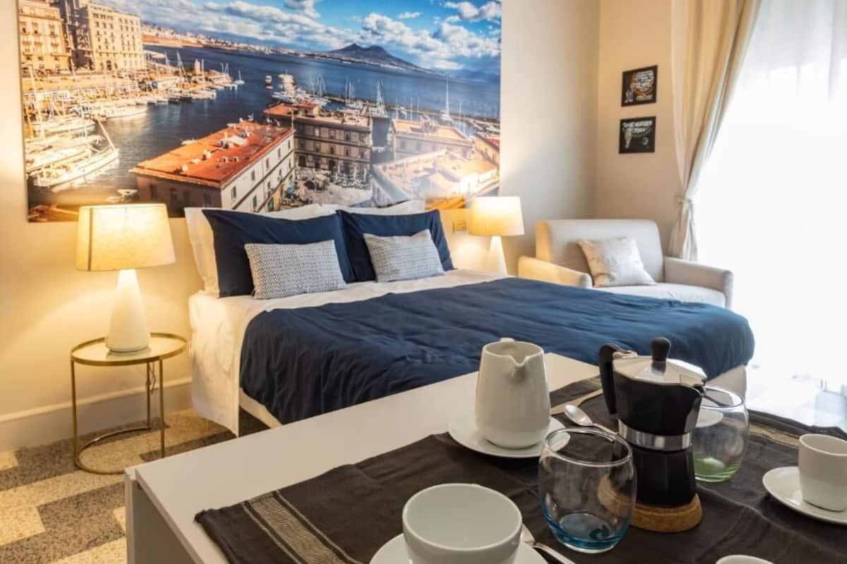 Ett hotellrum med en säng, ett soffbord med tekanna och tekoppar, en stol och en stor målning av en stadsbild på väggen.