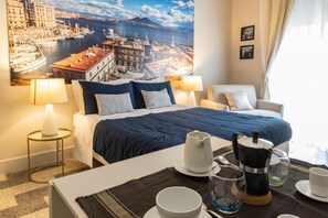 Habitación de hotel con cama, una mesa de centro con juego de té, una silla y un cuadro grande con una vista urbana.