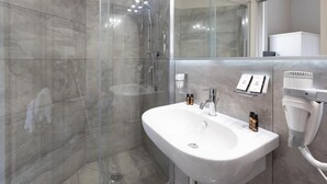 A modern bathroom with a glass-enclosed shower, a white sink with two bottles of shampoo, and a hairdryer mounted on the wall.