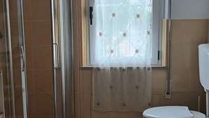 Une salle de bain comprenant des toilettes, un espace douche délimité par des portes en verre et une fenêtre agrémentée d’un rideau à motifs floraux.