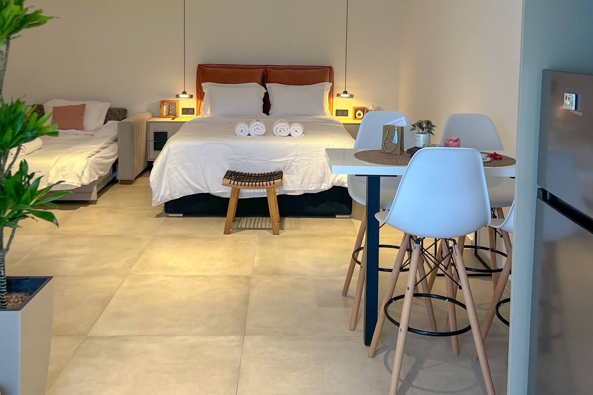 Ein modernes Schlafzimmer mit einem großen Bett, Nachttischen, einem kleinen Hocker, einem Esstisch mit Stühlen und einem Kühlschrank.