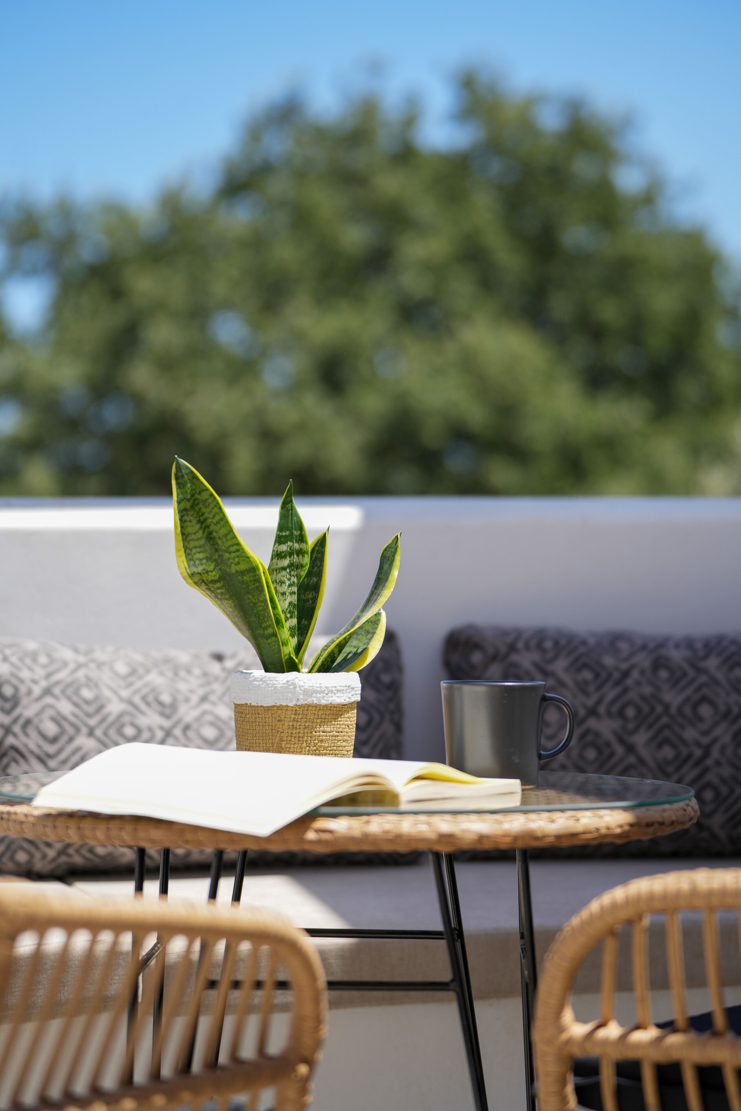Une chaise en osier, une petite table avec un livre et une tasse, et une plante en pot sur un balcon.