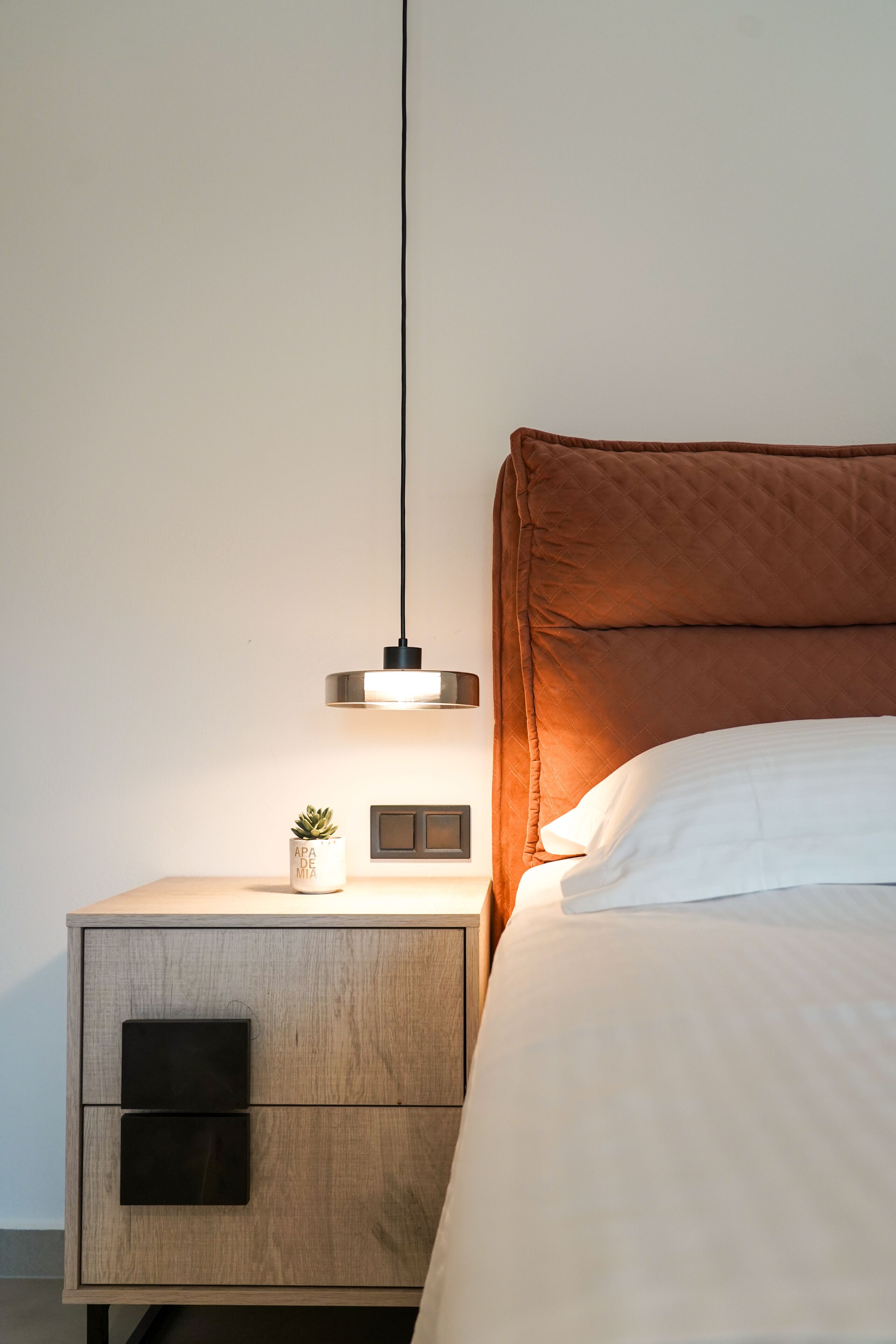 Una cama bien hecha con cabecera, una mesita de noche con lámpara y una pequeña planta en maceta.