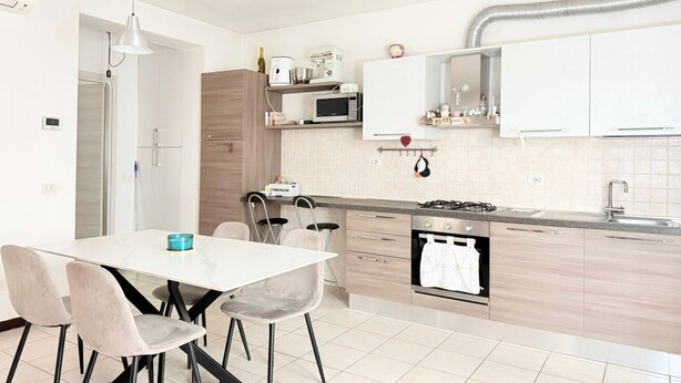 Cocina moderna con armarios blancos, microondas, estufa a gas y lavaplatos. Hay una mesa de comedor con sillas y una lámpara colgante.