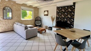 A modern living room with a stone wall, a grey sofa, a wooden coffee table, a dining table with chairs, and a large window.