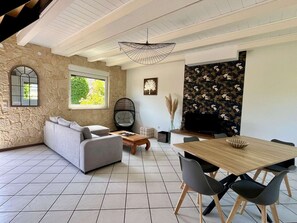 A modern living room with a stone wall, a grey sofa, a wooden coffee table, a dining table with chairs, and a large window.