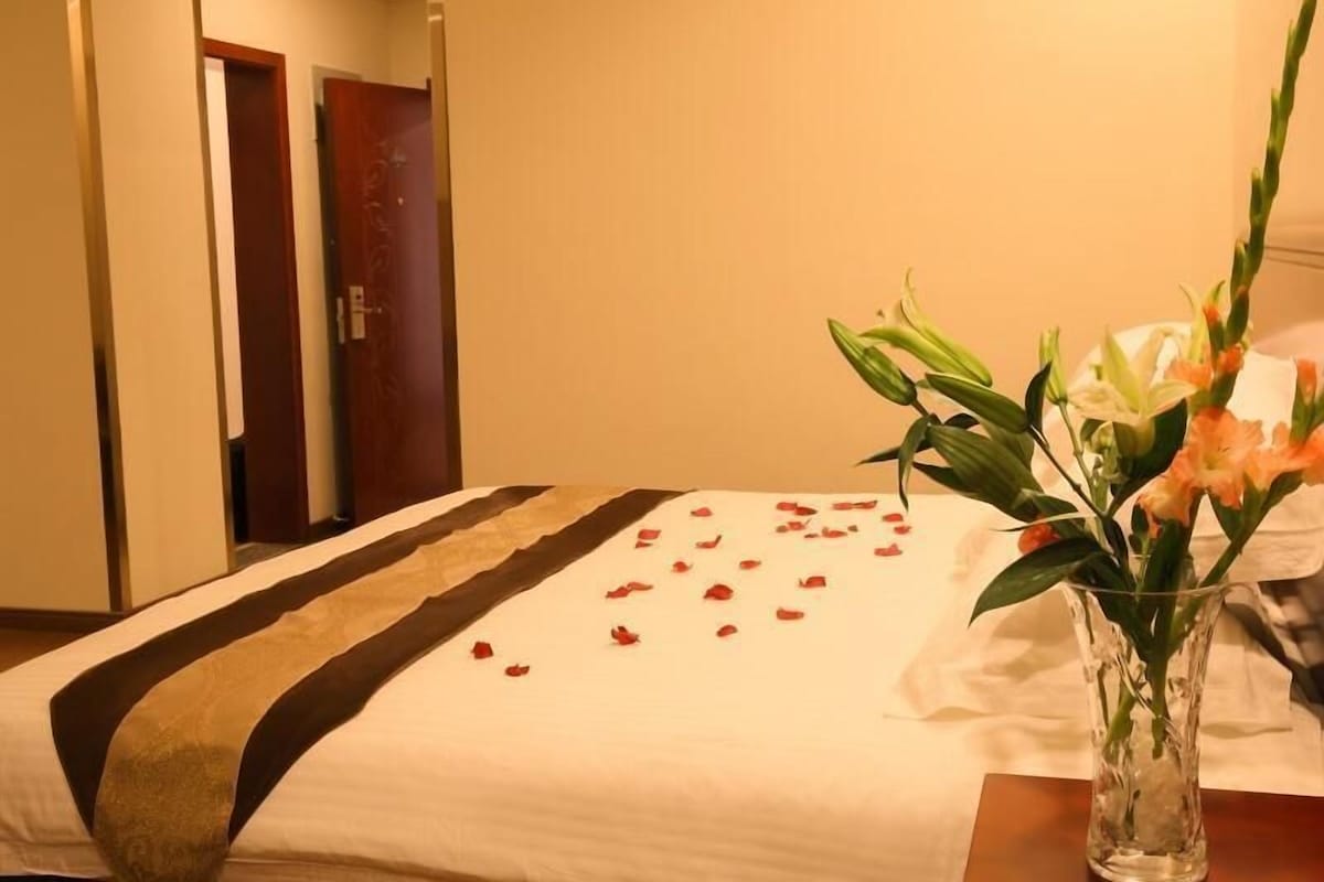 A hotel room with a bed, a vase with flowers, and a door in the background.
