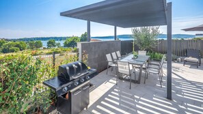 Eine Terrasss mit Grill, Esstisch, Stühlen und Blick auf einen See und Hügel.