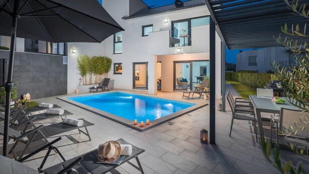 Zona de piscina exterior moderna amb gandules, zona de seients coberta i interior ben il·luminat.