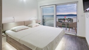 Un dormitori amb un llit gran, una nitu, una porta corredissa de vidre que dóna a un balcó amb taula i cadires, i vista al mar.