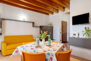 Zona pranzo con tavolo apparecchiato per due, divano e angolo cottura con televisore.