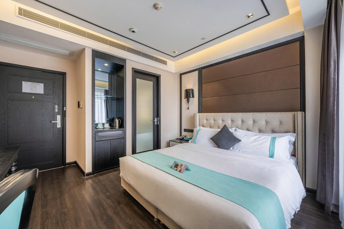 A hotel room with a large bed, a dark door, a mirror, and a bedside table with a lamp.