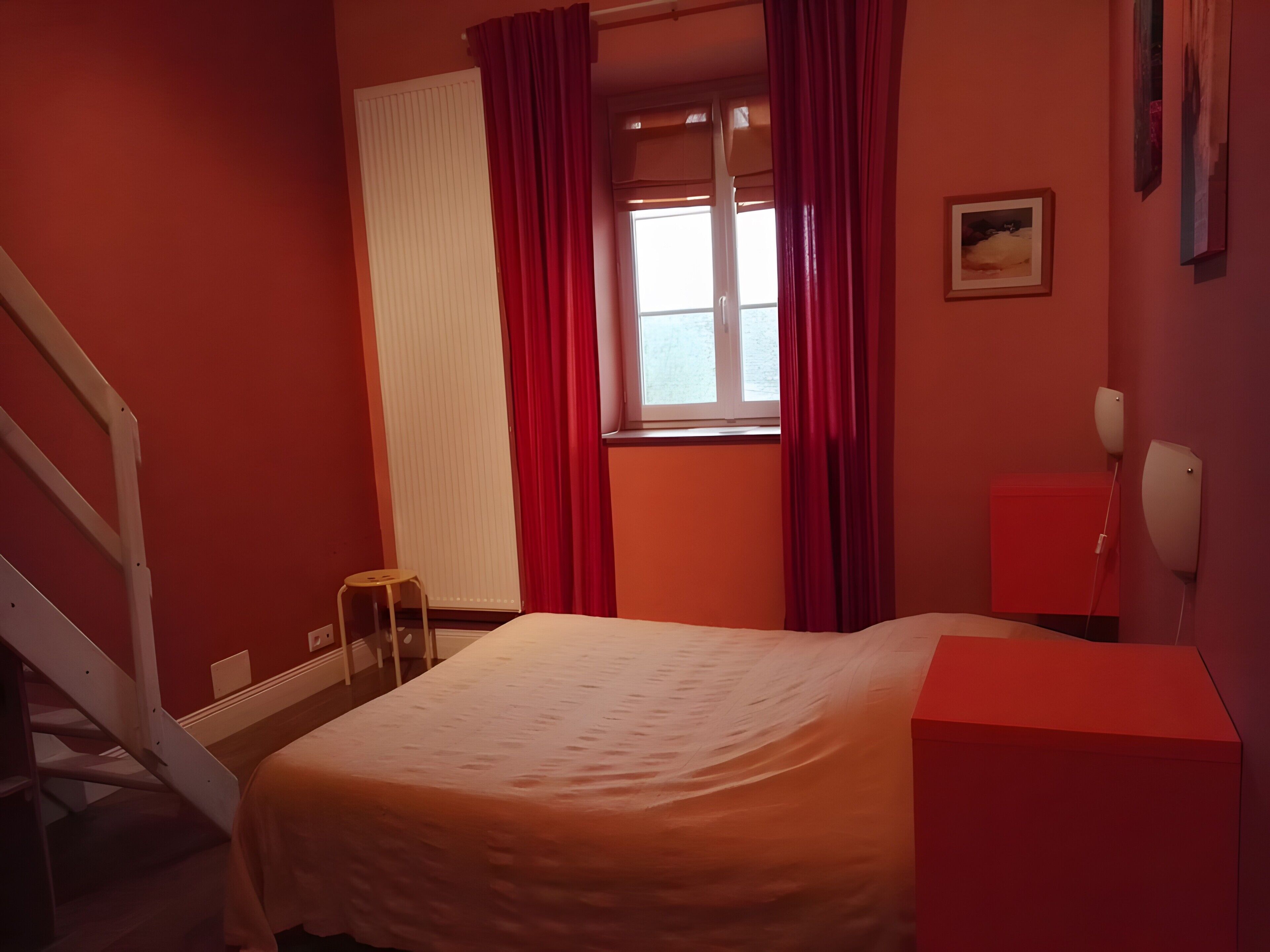 A bedroom with a bed, a window with curtains, a bedside table, a lamp, and a red wall.
