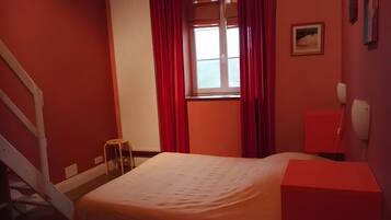 A bedroom with a bed, a window with curtains, a bedside table, a lamp, and a red wall.