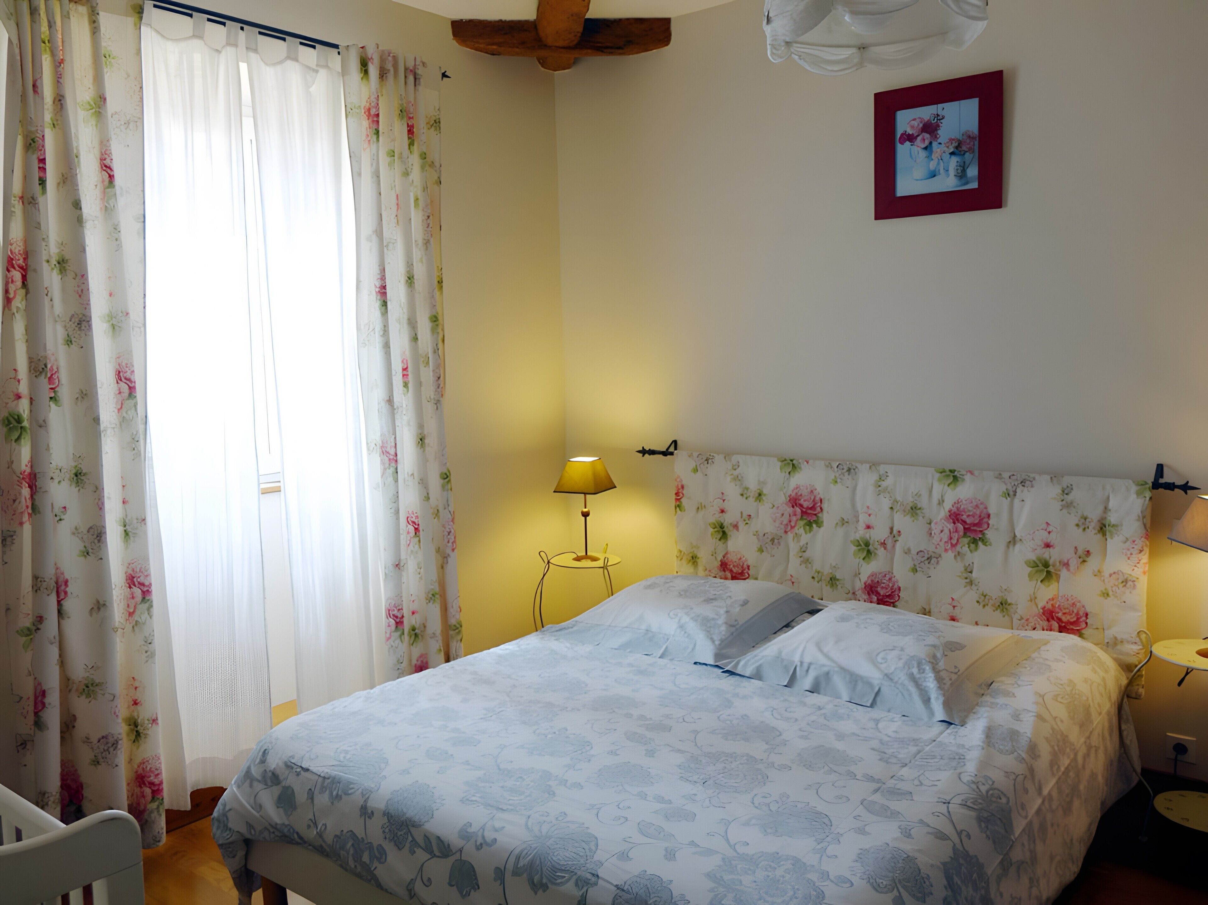 Une chambre à coucher avec un lit, des rideaux à motifs floraux, une lampe de chevet et un tableau encadré au mur.