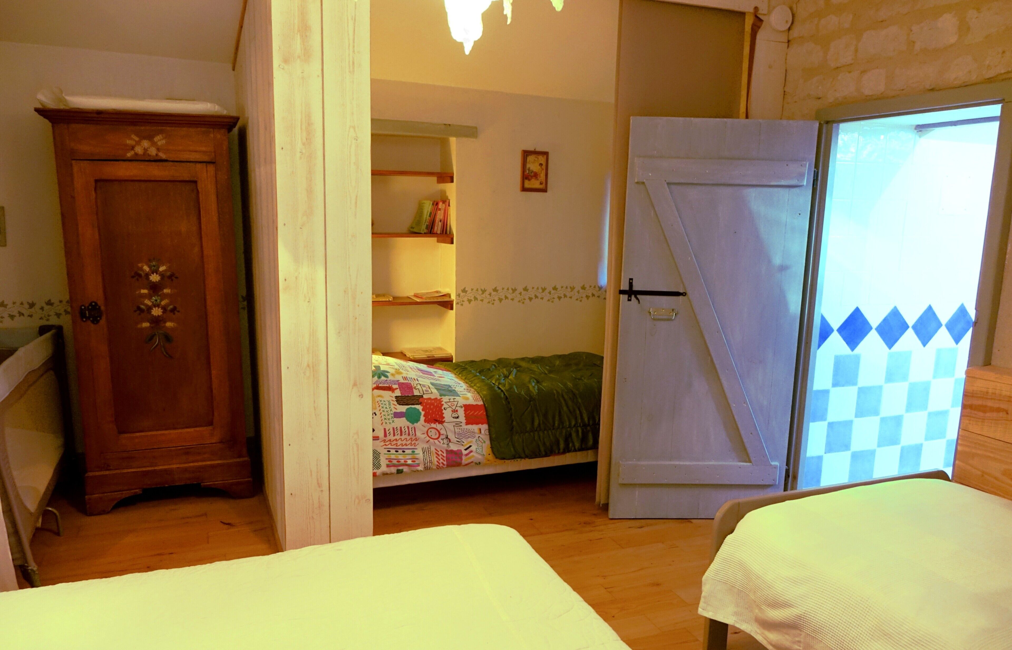 A bedroom with a wooden wardrobe, a bed with a colorful quilt, a small shelf with books, and a door leading to another room.
