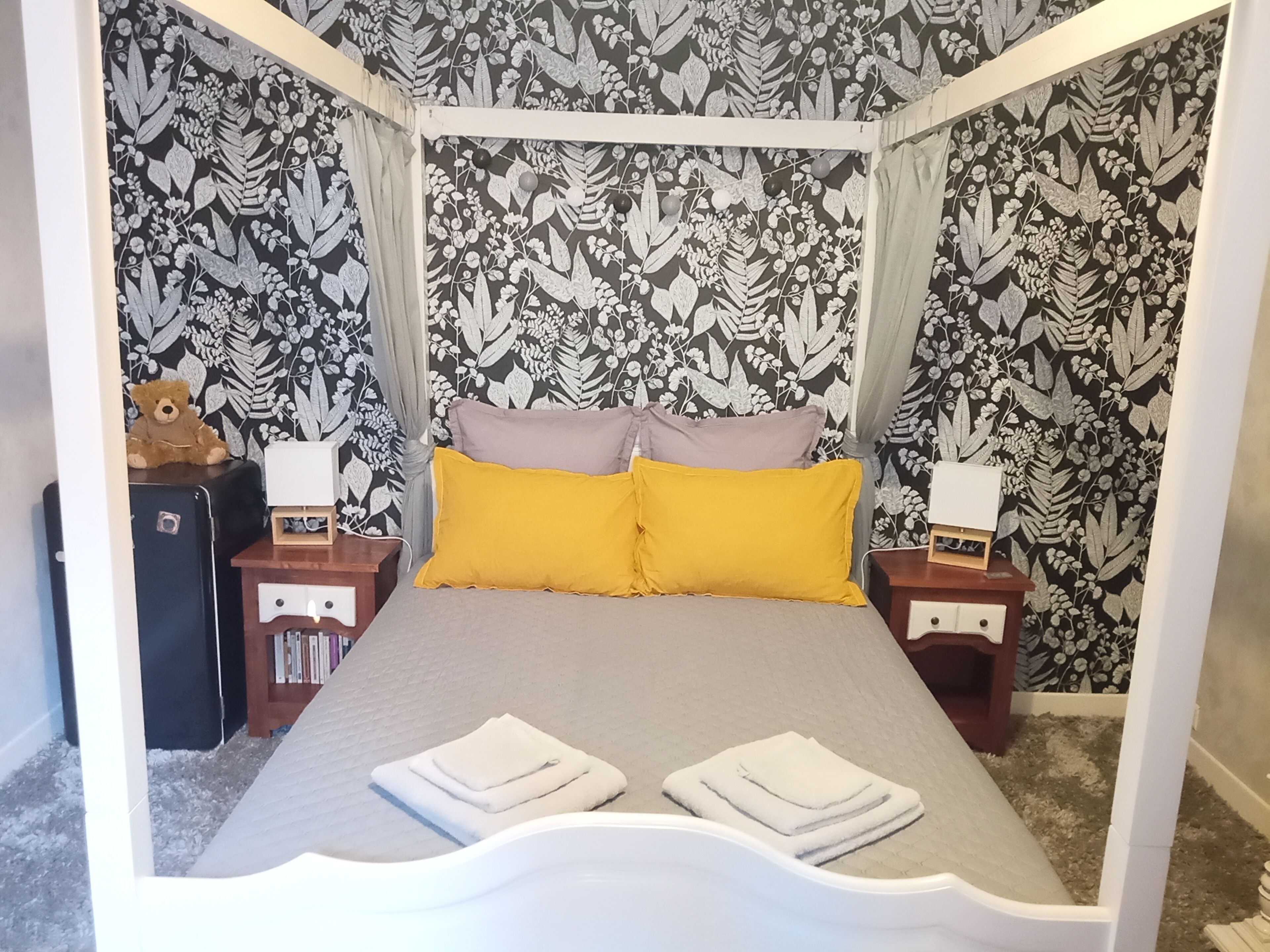 Una cama con dosel sostenida por cuatro pilares, con colcha gris y dos almohadas, una amarilla y otra gris, flanqueada por dos mesitas de noche con lámparas.