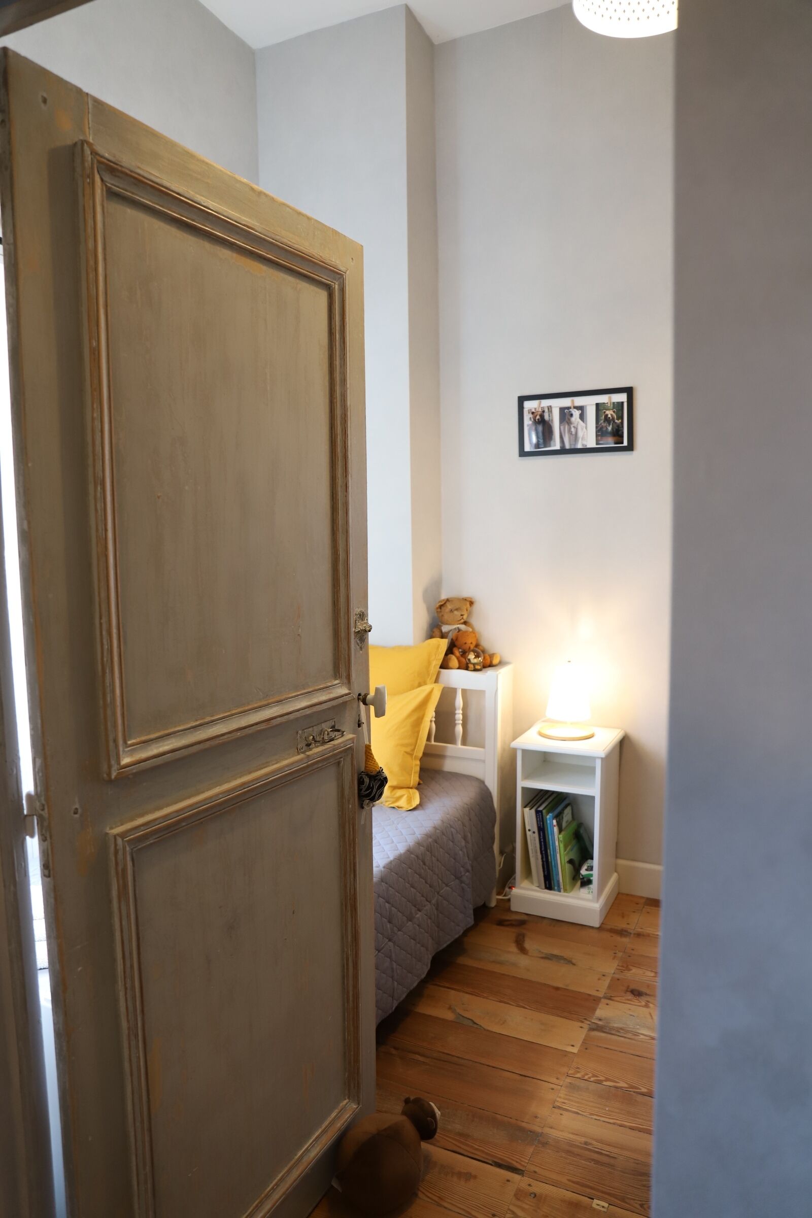 Una stanza con un armadio in legno, un letto con cuscino giallo, un comodino con lampada e una piccola mensola con libri.