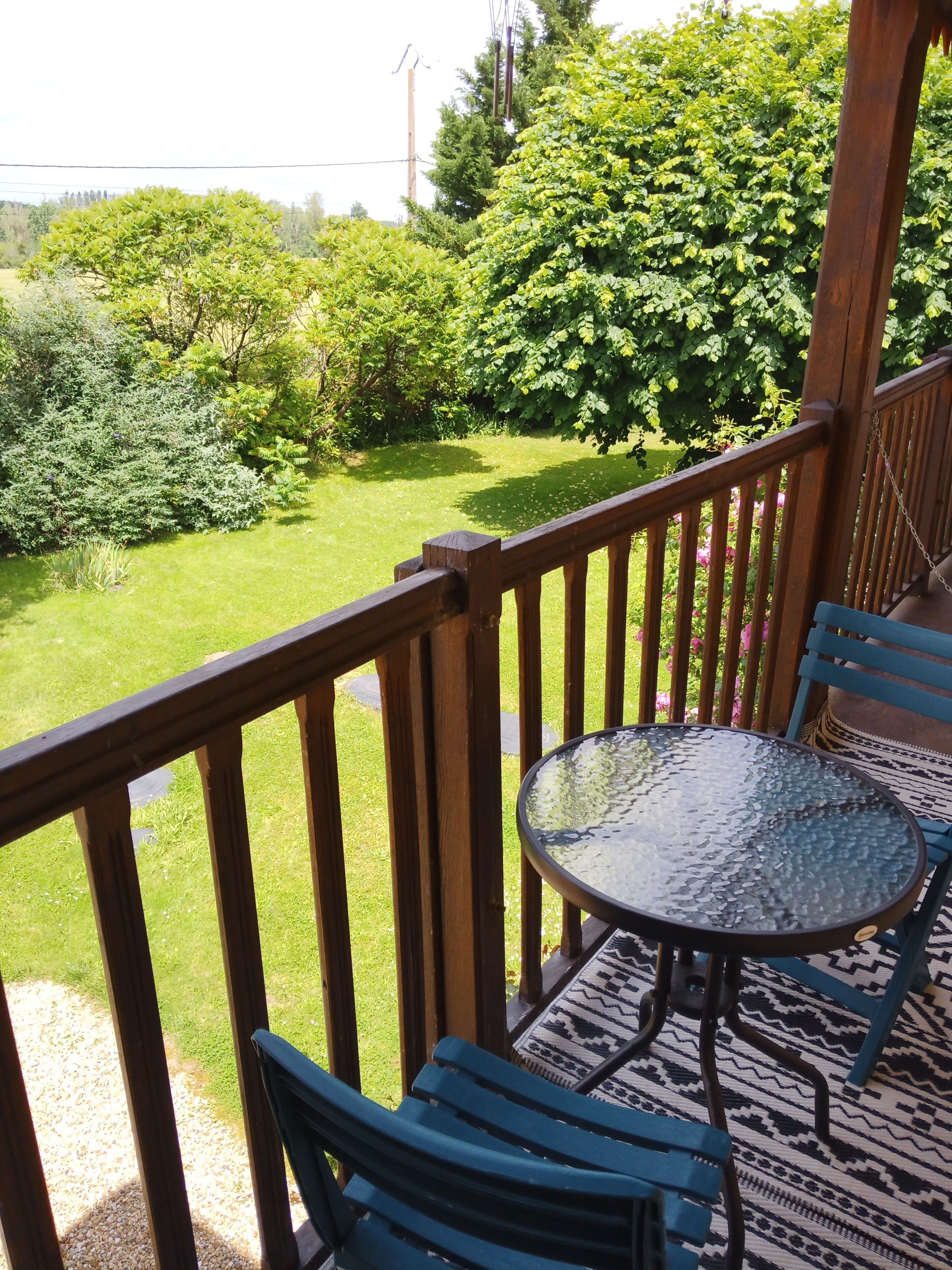 A wooden deck with a glass table and blue chairs, overlooking a well-maintained lawn and dense greenery.