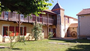 A courtyard with a building featuring a tower, wooden balconies, and a stone wall with a large wheel.