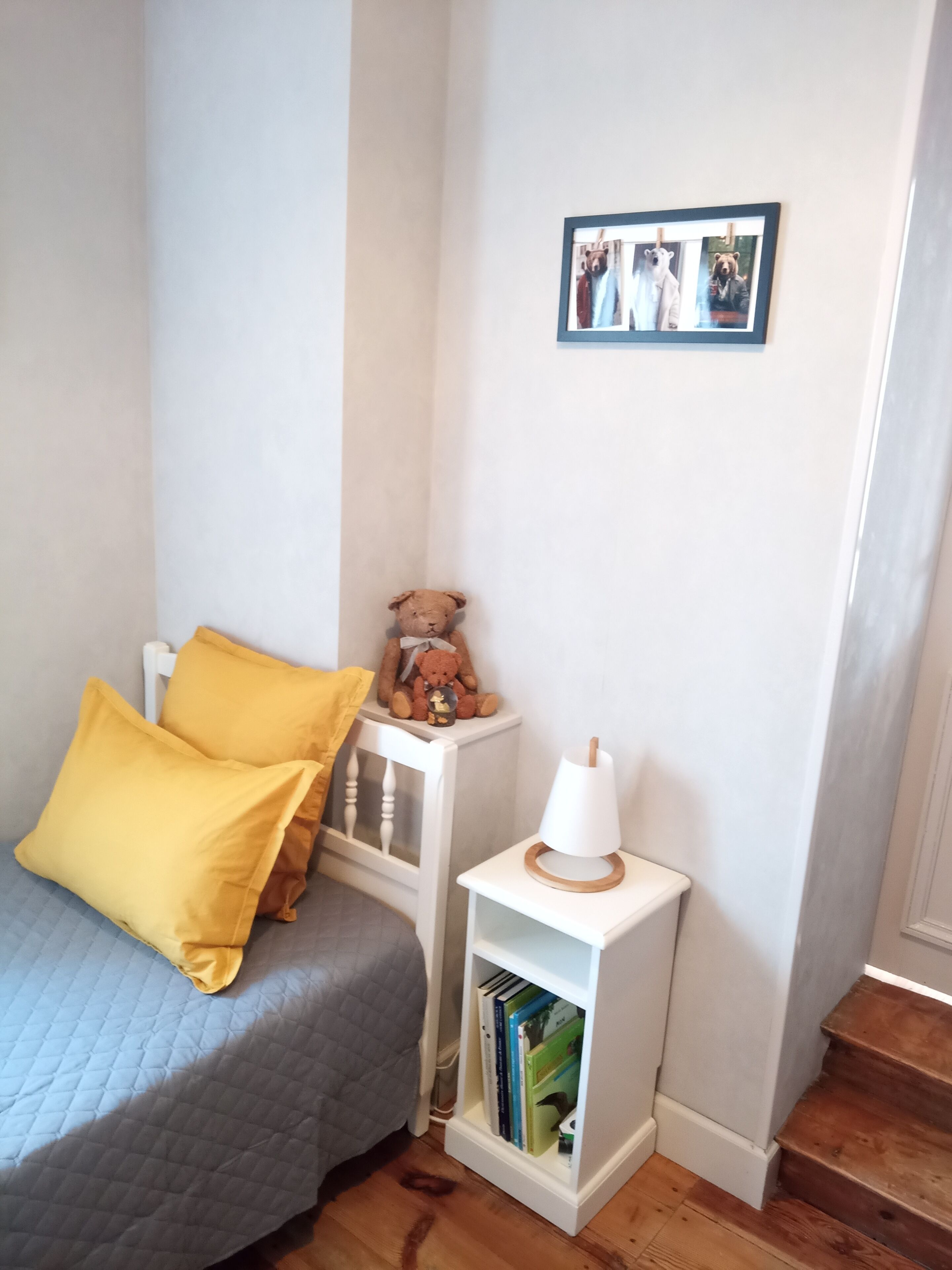 A bedroom with a bed, a small bedside table with a lamp, a bookshelf, and a teddy bear.