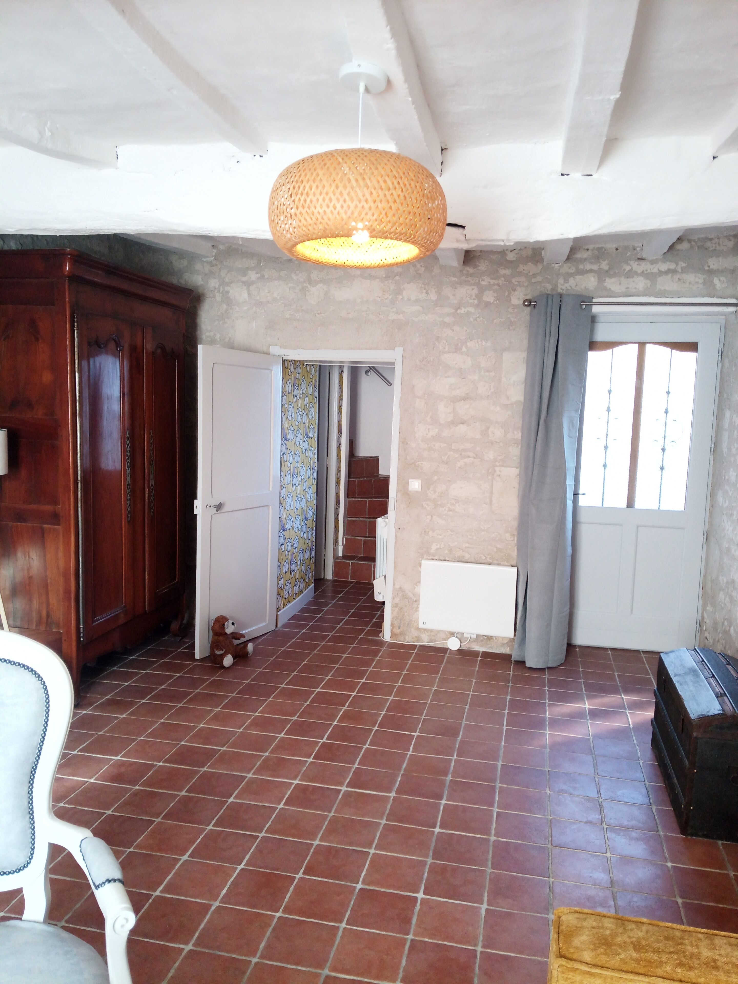 A spacious room with a tiled floor, a wooden wardrobe, a white chair, a wicker pendant light, and a door leading to a staircase.