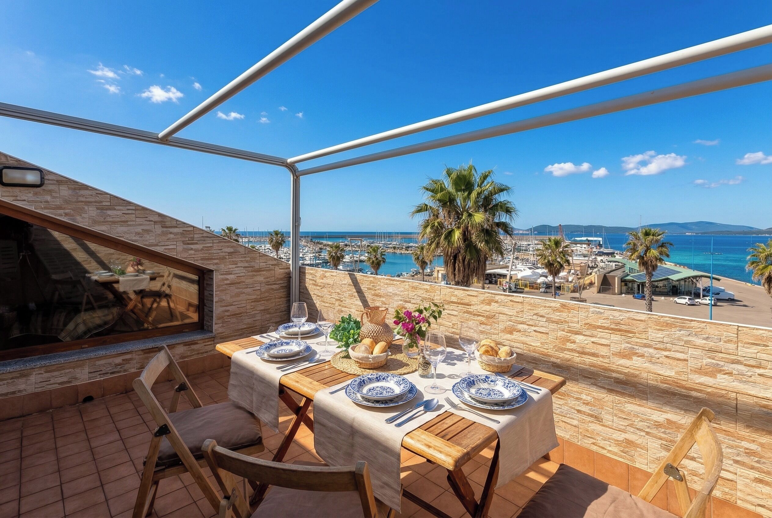 A terrace with a table set for dining, overlooking a coastal area with palm trees and a view of the sea.