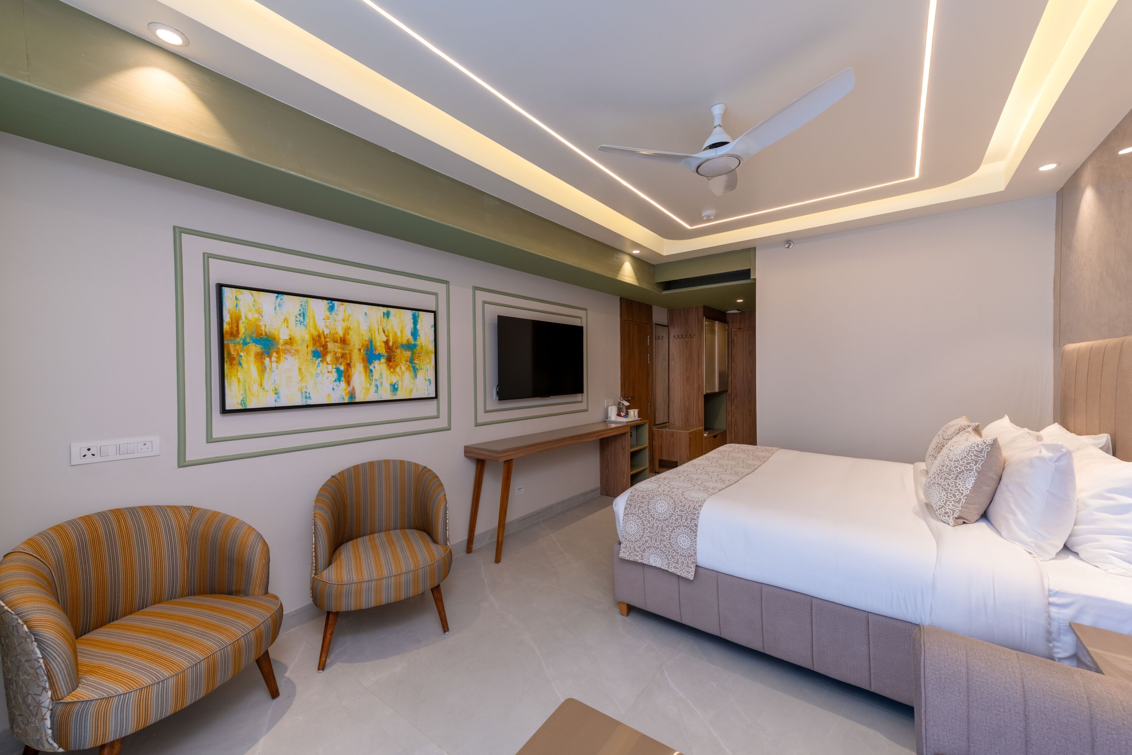 A modern bedroom with a bed, two armchairs, a TV, and artwork on the wall.