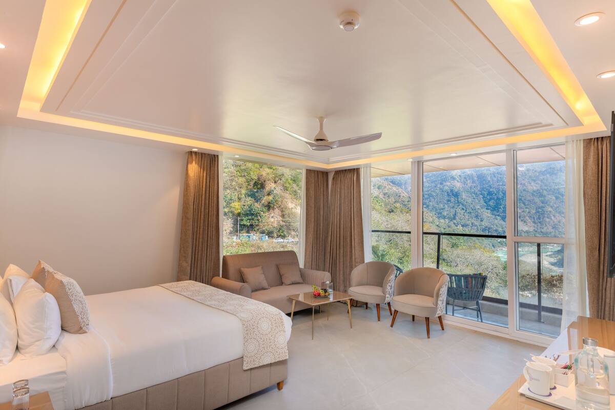 Una habitación de hotel moderna con una cama grande, un sofá, una mesa de comedor y una silla. Cuenta con un balcón con vistas a las montañas.