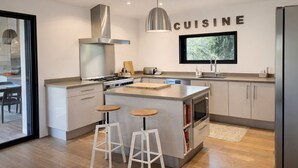 Een moderne keuken met een centraal eiland, barstoelen, een roestvrijstalen afzuigkap en een bordje met daarop 'CUISINE'.