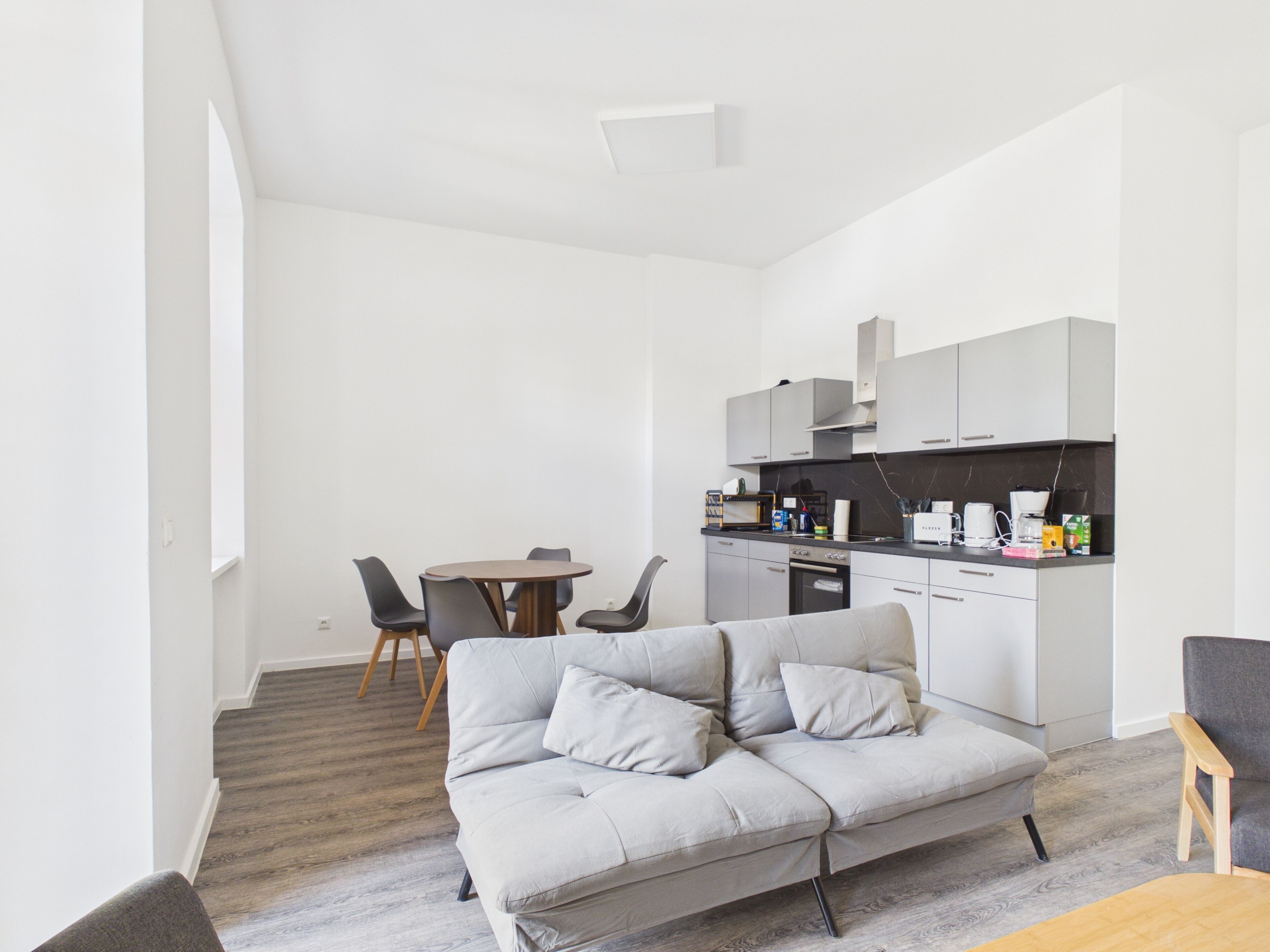 Ein modernes Wohnzimmer mit grauem Sofa, einem Esstisch mit Stühlen und einer Küchenzeile mit Schränken und Geräten.