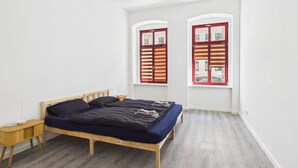 Une chambre à coucher avec un lit en bois, une table de chevet, deux fenêtres avec des volets rouges et un mur blanc.