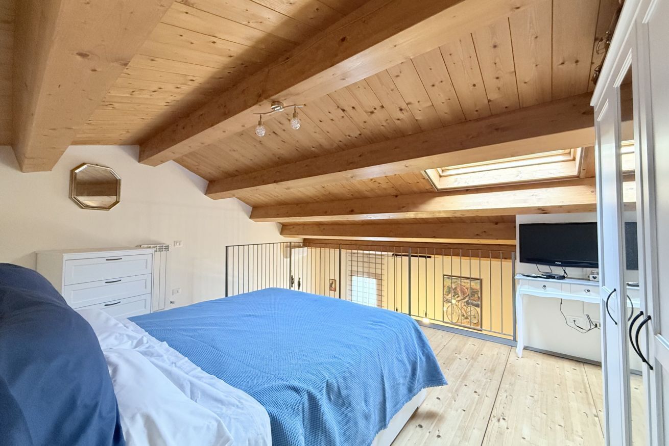 Quarto com cama, escrivaninha com televisão, claraboia e varanda com bicicleta.