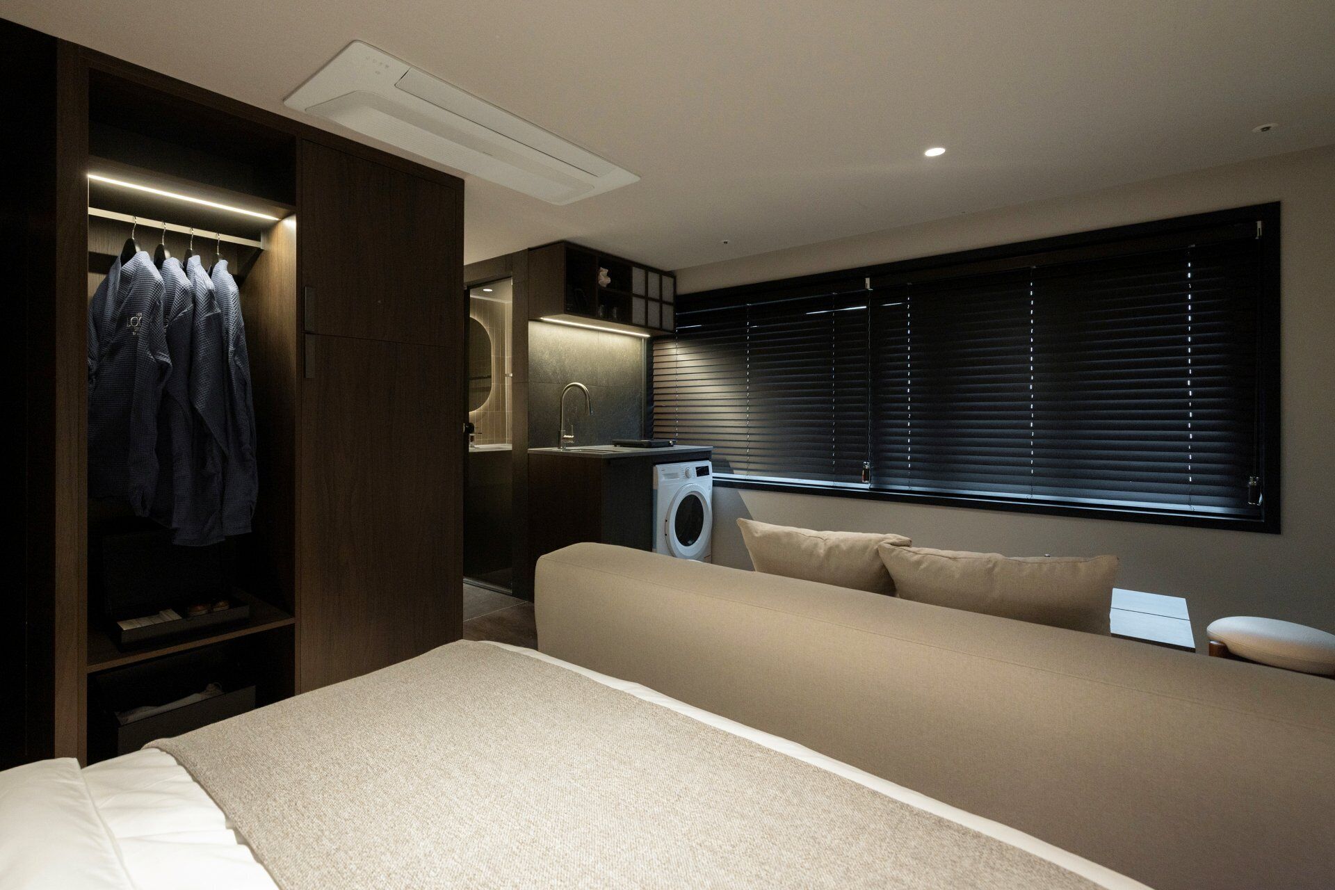 Quarto compacto com cama, máquina de lavar embutida, armário e janela com persianas.