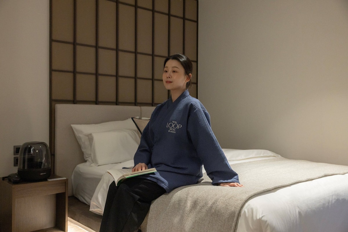 Eine Person sitzt auf einem Bett in einem Hotelzimmer mit Blick auf eine Stadtlandschaft durch ein Fenster.