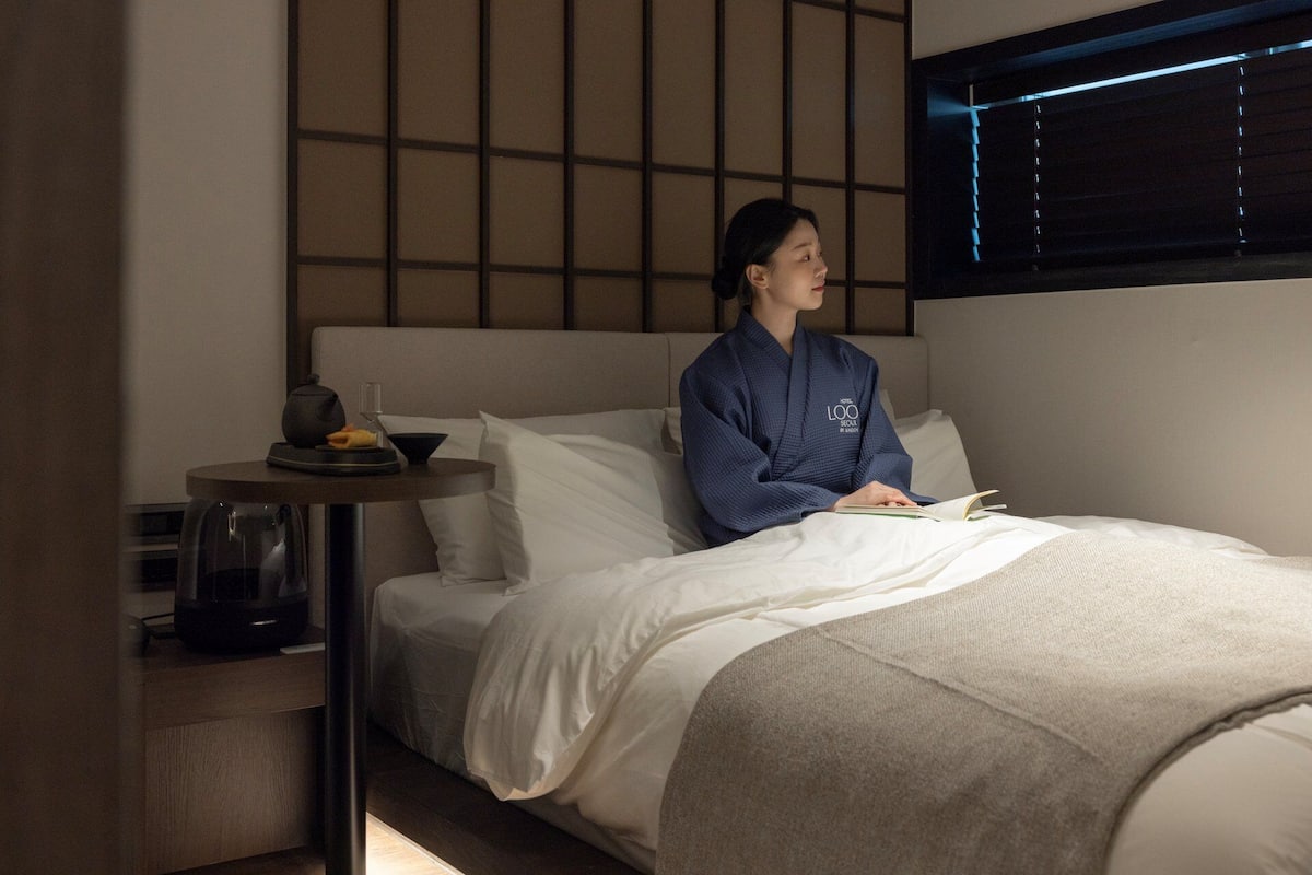 Ein Hotelzimmer mit einem Bett, einem Nachttisch mit Lampe und einem Fenster mit Jalousien.