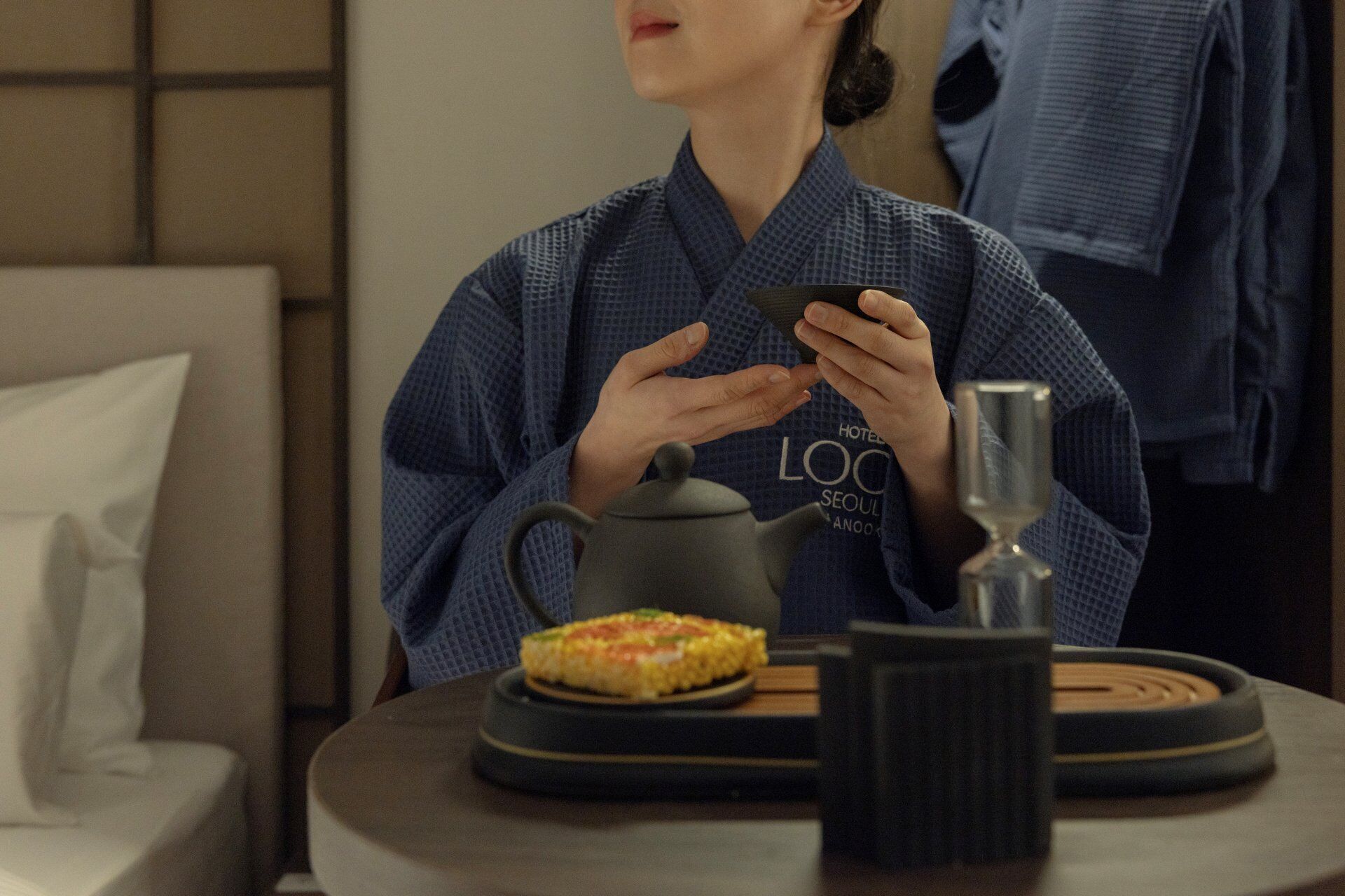 A person in a blue robe sitting at a table with a teapot, a glass, and a plate of food.