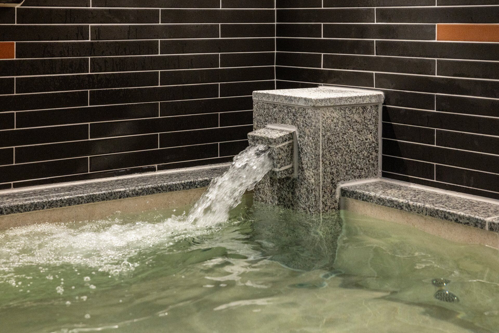 A stone hot tub with water flowing into it, set against a tiled wall.