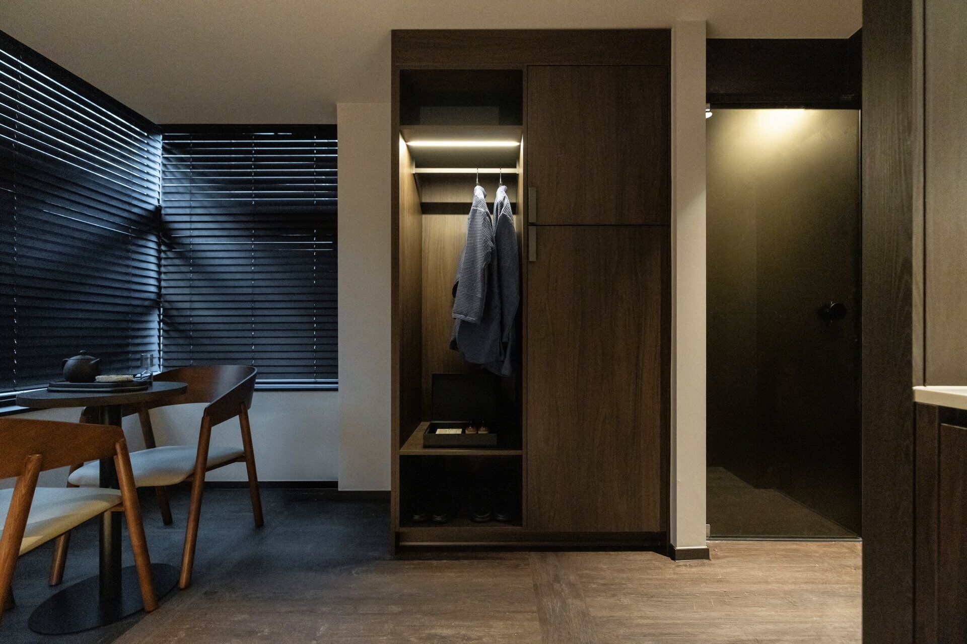 A modern room with a wooden floor, a desk with a chair, a window with blinds, and a built-in wardrobe with hanging clothes.