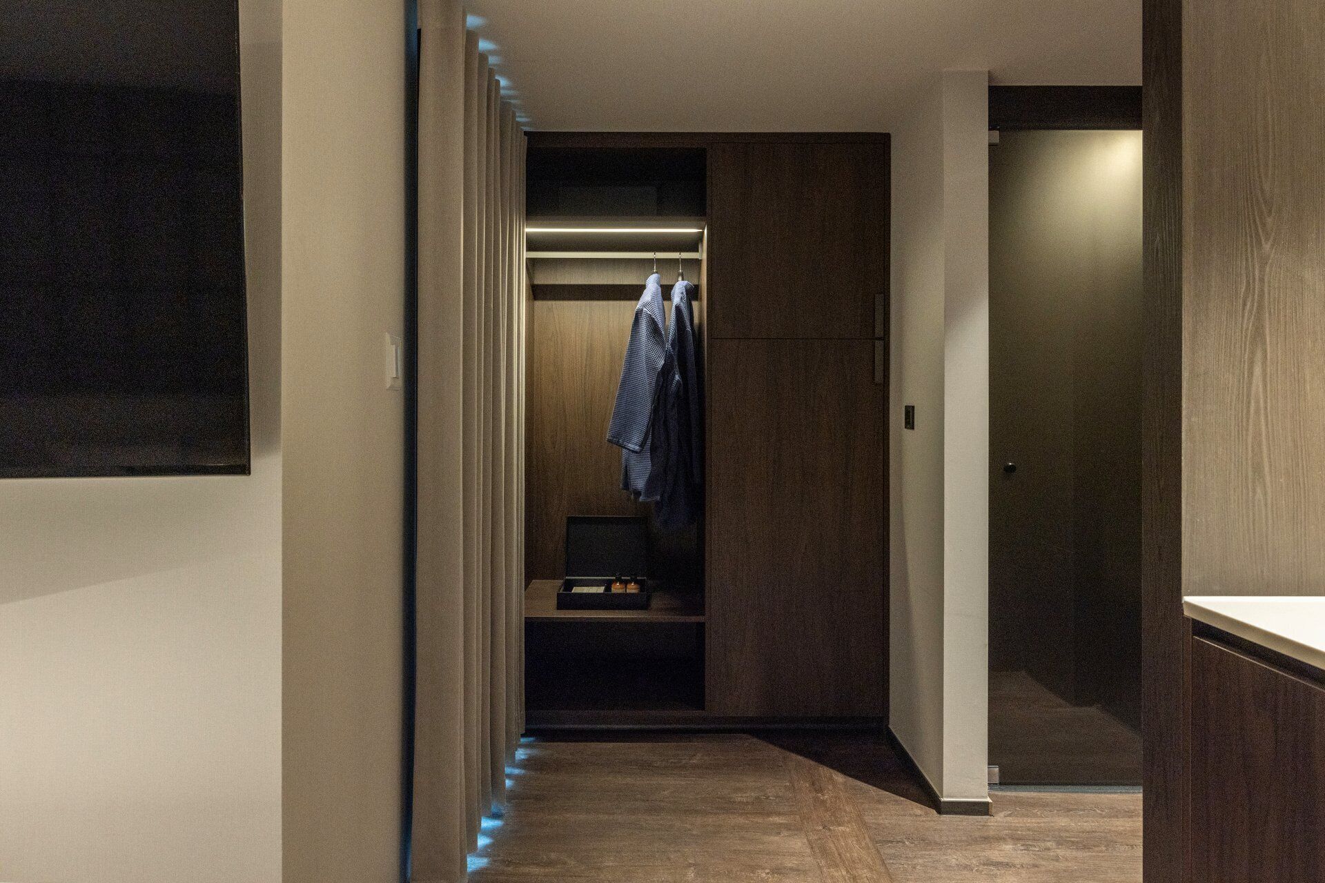 A narrow hallway with a wooden floor, a closet with hanging clothes, and a door on the right.