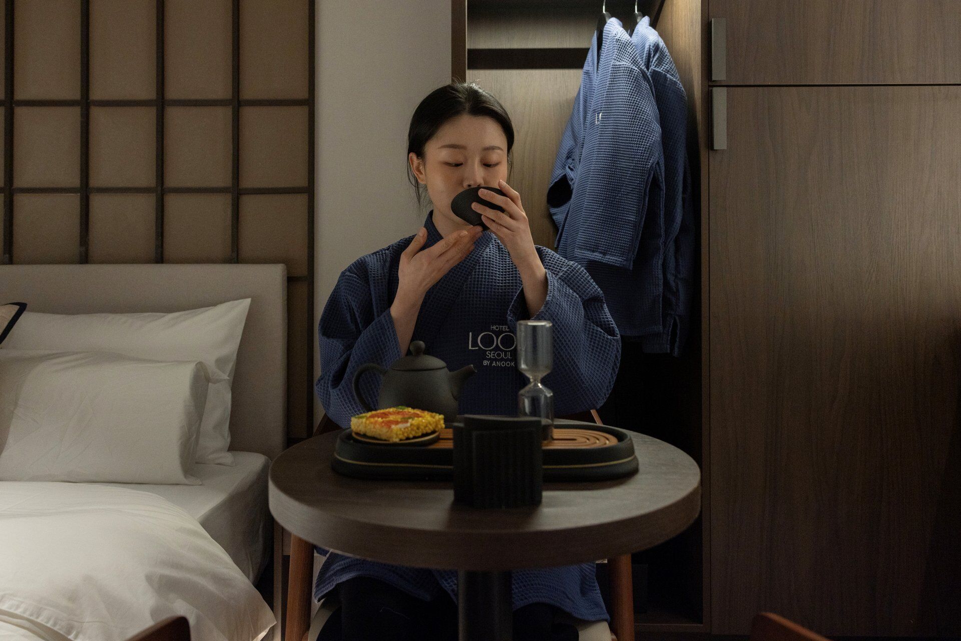 A person seated at a round table with a teapot, a glass, and a plate of food in a hotel room.