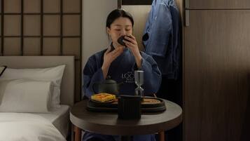 A person seated at a round table with a teapot, a glass, and a plate of food in a hotel room.